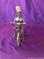 Front view of silver metal figurine of a person riding a bicycle wearing top hat and glasses