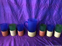 Full set of one blue plastic pitcher and five plastic cups, each with a woven straw base, displayed against purple background.