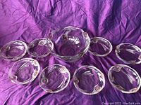 One large clear glass bowl surrounded by eight matching small dessert plates, all having wavy rim designs, placed on purple cloth.