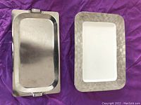 Two rectangular serving trays side by side on a purple cloth. Left tray is solid silver-tone metal with handles, right tray is white center with textured silver-tone border.