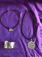 Two sterling silver necklaces with black leather cords and one cuff bangle bracelet arranged on purple background.
