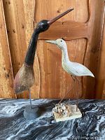 Two bird carvings on a wood table with a wood paneled wall background; one black and brown tall with a slender neck, the other white on metal legs with a decorated base.