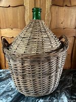 Large wicker-wrapped glass demijohn with conical wicker top and two side handles shown from the front on a marble surface with wooden cabinet background.