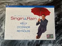 Front cover of Singin' in the Rain 60th Anniversary Ultimate Collector's Edition Blu-Ray/DVD boxed set showing Gene Kelly holding umbrella in rain motif.