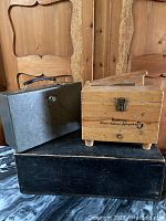 Photo showing all three boxes stacked: light wood shoeshine box on top, gray metal cash box in middle, black wooden box at bottom.