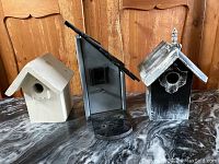 Photo of three different styled wood birdhouses including a cream wooden, gray metal, and black rustic birdhouse on a marble surface in front of wooden background.