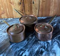 Three copper pots with lids arranged on a marble surface in front of a wooden wall. All have long metal handles and copper lids.