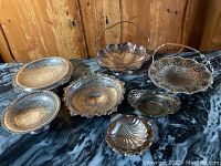Seven vintage silver plate decorative items arranged on a dark marble surface, showing variety of trays, baskets, and dishes with ornate engraving and pierced edges.