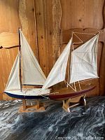 Photo showing both wooden sailing boat models side by side on display stands against wooden wall background.