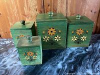 Set of four vintage green wooden canisters arranged in a row, showing front floral designs and top knob lids.