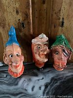 Three vintage large plastic puppet heads showing varying colored hats and detailed facial features against wooden background.