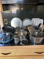 Overview of all bakeware and utensils including cake tins, springform pan, muffin tins, bundt pan, graters, potato masher, and glass pie plate arranged on countertop.