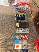 Stack of board games including Hedbanz, Connect 4, Jeopardy Junior, Guesstures, Scrabble, Monopoly, domino set in wooden box, and 500-piece cat puzzle box.