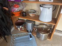 Wide shot showing multiple kitchen items including enamel coffee pot, stock pot, colander, French press, and dish drying rack on wooden shelving unit.