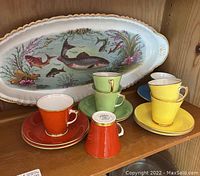 Porcelain fish oval platter and demitasse cups in orange, yellow, green, and blue with saucers arranged on a wooden shelf