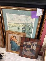 Three framed prints leaning against a wall: a large seascape with boats, small portrait of a woman and child, and another small print showing an interior scene. The largest frame is wood with a golden inner edge.