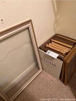 Photo showing a large beige rectangular empty picture frame leaning against a wall with other frames stacked in a box beside it.