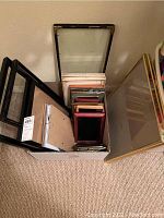 Photo showing multiple assorted picture frames stacked within a box, including various sizes and styles with visible glass panels and backing.