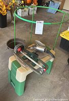 Garden bench with tools including hand trowels, sprinklers, bucket, and green circular plant support frame in garage setting