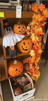 Photo shows Halloween pumpkin decorations including three pumpkins with carved faces, a small ghost figure in pilgrim hat, and a cardboard box with assorted small decor items on a shelf. Orange and yellow fall leaf garland hangs to the right side.