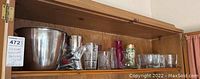 Wide view of the top shelf of wooden cabinet holding the items. From left to right: vintage metal ice bucket, a plastic bag with cocktail supplies, etched and textured drinking glasses, 12 small vases of varying styles and colors including pink and amber glass.