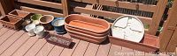 All planters and pots arranged on wooden deck, showing variety of shapes and sizes including rectangular and round forms in ceramic and plastic.
