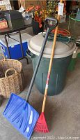 Photo showing blue snow shovel with black handle standing next to a green Rubbermaid trash can with lid and red garden rake with wooden handle.
