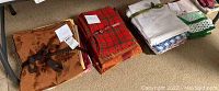 Photo showing three stacks of table linens: brown floral-patterned linen, red plaid linen, and a stack of white linens with various trims.