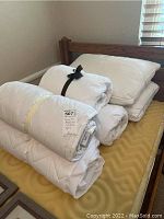 White twin bedding items stacked on a twin mattress including mattress pads, bedspreads tied with ribbons, and four pillows.