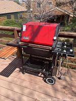 Photo of Weber grill on wooden deck showing full grill with red lid, black frame, wheels, wooden side table and metal side control panel with knobs and temperature gauge