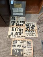 Photo of multiple Boston Globe newspapers with large headlines about end of war in Europe and V-E Day, along with a framed memorabilia piece containing clippings and typed notes.