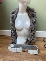 Fur shawl draped on a white mannequin torso, white leather belt placed below the shawl on the same surface.