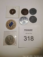 Seven coins laid out on a white background: includes Calgary Stampede 1978 token, Apollo 11 commemorative coin, small 'T' coin, two irregular crossed coins, silver coin in sleeve, and detailed engraved coin.