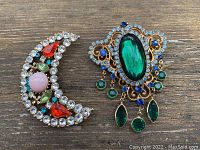 Photo showing two rhinestone brooches on wood surface: crescent moon shape with multicolored stones, and ornate brooch with green central stone and dangling green and blue stones.
