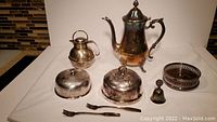 Lot arranged on table showing a large teapot, smaller teapot, bell, two bottle coasters, two cocktail forks