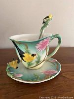Side view of painted porcelain teacup, saucer, and spoon with bird and floral design