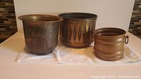 Three vintage brass and copper plant pots of varying sizes side by side on white cloth.