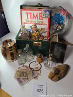 Photograph showing entire green vintage case open with all contents including magazine, glass bottles, banknotes, wooden item, and other collectibles laid out.