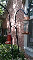 View of two black metal plant hangers; one holds a copper and glass bird feeder and the other a red planter