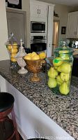 Wide view of kitchen counter holding two glass canisters with faux pears, a footed amber glass bowl with faux lemons, and a ribbed glass urn with faux lemons.