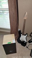 Photo of Pearl Cajon with black front and natural wood sides, next to Fender Starcaster electric guitar leaning against wall near window.