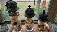 Three graduated glass canisters with dark ornate metal lids on windowsill filled with decorative spheres.
