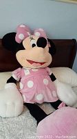 Front view of large Minnie Mouse plush toy seated on a bed, showing pink dress with white polka dots and matching bow, with tags attached.