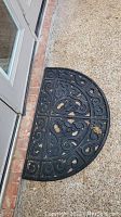 One black semi-circular rubber door mat with decorative scroll pattern placed outside a door on a textured floor, showing signs of debris and weathering.