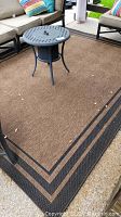 Brown outdoor rug with a black and dark brown patterned border laid out under patio furniture on a concrete surface.