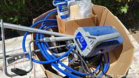 Full view of Graco Magnum paint sprayer device with hose and handle inside a cardboard box outdoors.