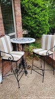 Full view of outdoor round patio table with mosaic tile top and two matching metal bar height chairs with striped cushions.