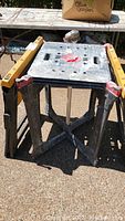 Pair of black plastic sawhorses and work stand with yellow tool pouch on outdoor surface