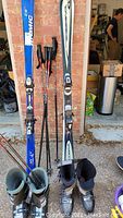 Image showing two pairs of skis (blue Atomic and silver-white Cobra), three pairs of ski poles, and two pairs of ski boots placed on ground