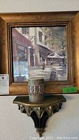 Framed print with wooden frame depicting outdoor cafe street scene, with metal wall sconce shelf below holding candle with metal filigree holder.